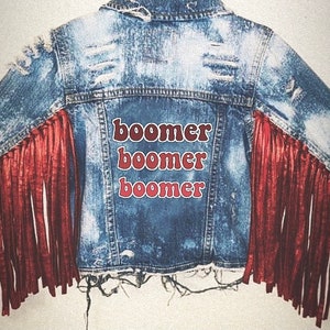 May include: A blue denim jacket with red fringe on the sleeves. The back of the jacket has the word "boomer" written three times in red.