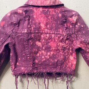 May include: A purple denim jacket with bleach splatters and distressed edges. The jacket has a classic jean jacket style with a button closure and pockets.