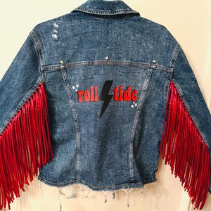 May include: A blue denim jacket with red fringe on the sleeves. The back of the jacket has a black lightning bolt and the words "roll tide" in red.