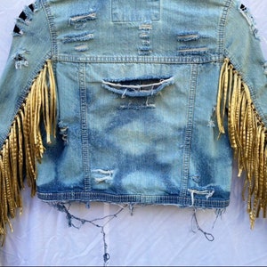 May include: A blue denim jacket with gold fringe trim on the sleeves. The jacket has distressed details and a cropped fit.