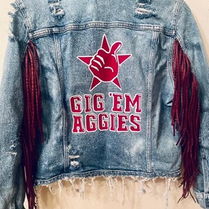 May include: A blue denim jacket with maroon fringe on the sleeves. The back of the jacket has a red star with a white hand giving a thumbs up and the text "Gig 'Em Aggies" in white letters.