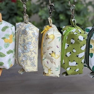 Puede incluir: Seis pequeñas bolsas con cremallera con diferentes patrones de tela. Las bolsas están colgadas de anillos metálicos y tienen un tirador de cremallera amarillo. Los patrones incluyen diseños florales, animales y geométricos.