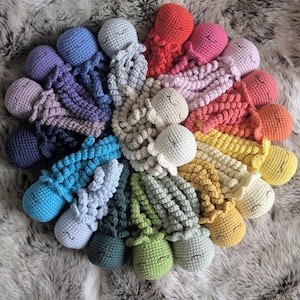 May include: A collection of small, handmade crochet octopus toys arranged in a circular pattern. Each octopus features a round head and eight curly, tentacle-like arms, with embroidered closed eyes and a simple smile on each face in various colours.