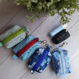 Op de afbeelding: Vier kleine ritsende tasjes met bloemenpatronen in tinten blauw, groen en rood. Elk tasje heeft een zilveren sleutelhanger. De tasjes liggen op een licht houten oppervlak.
