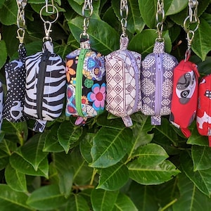 May include: Ten small zippered pouches with various patterns and colours, including cow print, zebra print, polka dots, floral, geometric, and a pug design. The pouches are attached to key rings and hanging from a metal rack.
