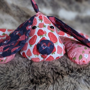 May include: A pink and blue fabric dog toy with a strawberry print. The toy has a black nose and black eyes.