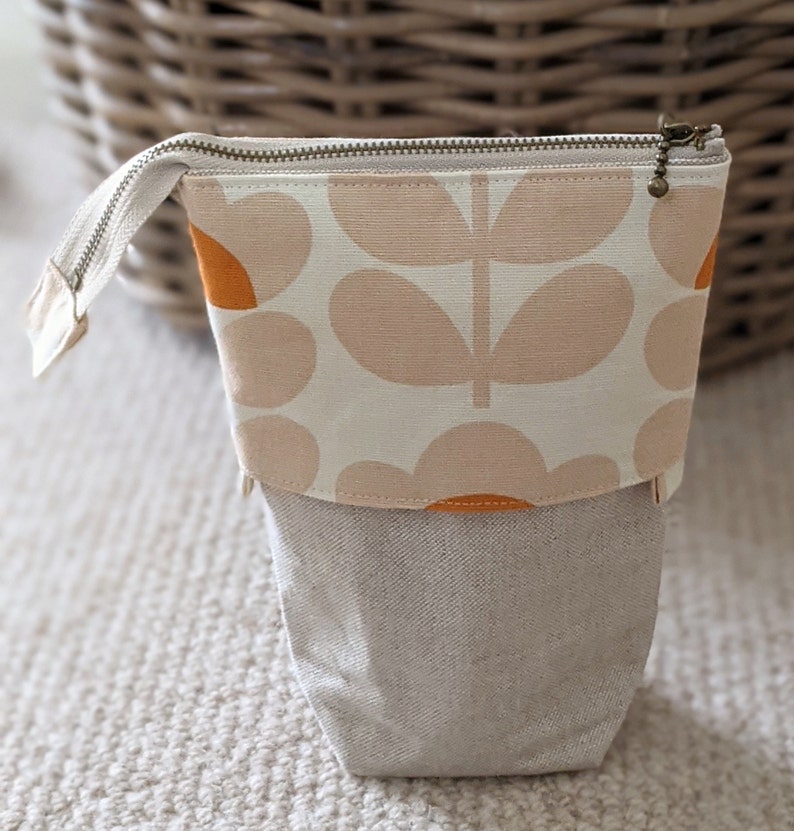 Puede incluir: Una bolsa de cremallera beige con un patr&oacute;n floral en naranja y beige. La bolsa est&aacute; hecha de tela y tiene una textura similar al lino.