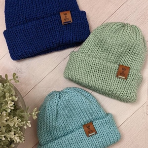 Peut inclure: Trois bonnets en tricot de différentes couleurs : bleu marine, vert clair et turquoise. Chaque bonnet est orné d’une petite étiquette en cuir marron sur laquelle est inscrit « Made by Nettie ».