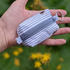 May include: A small grey and white striped zippered pouch with a bee design. The pouch has a silver key ring attached.
