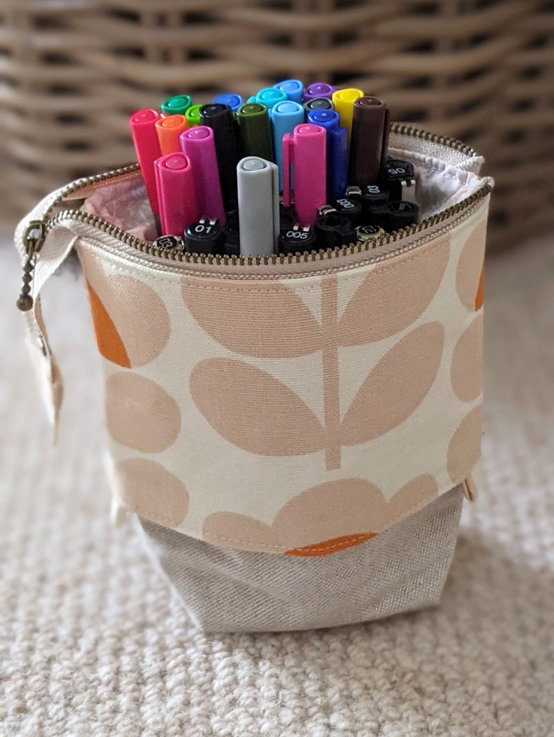 Puede incluir: Un estuche de l&aacute;pices de tela blanca con un patr&oacute;n floral en naranja y rosa. El estuche est&aacute; lleno de marcadores de varios colores.