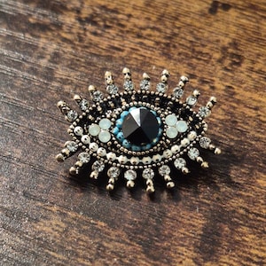 May include: A gold-toned evil eye charm with a black centre and white and blue accents. The charm is surrounded by a halo of clear rhinestones.