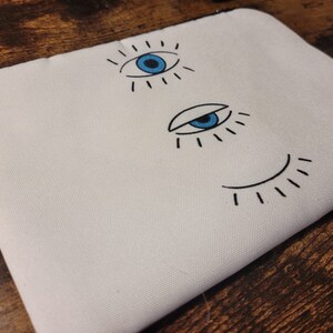 May include: White canvas pouch with a black and blue eye design. The eye is outlined in black and has a blue iris and pupil. The pouch has a zipper closure.