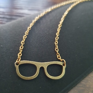 May include: A gold chain necklace with a pair of glasses pendant. The glasses are made of metal and have a simple, classic design.