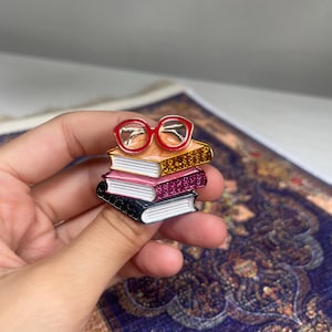 Könnte beinhalten: Ein Stapel von vier bunten Emaille-Büchern mit einer roten Brille oben. Die Bücher sind schwarz, rosa und goldfarben, mit weißen Seitenrändern. Die Brille hat einen goldenen Rahmen.