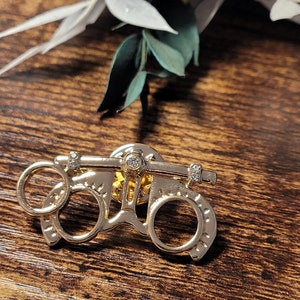 May include: Gold-toned eye chart pin with three small clear rhinestones. The pin is in the shape of an eye chart with two circles and a bar across the top.