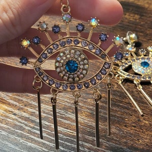 Pode incluir: Brincos dourados com um design de olho turco azul e correntes penduradas. O olho turco é rodeado por strass transparentes e roxos.