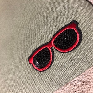 May include: Red embroidered sunglasses patch with black mesh detail on a green canvas background.