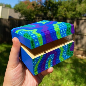 Puede incluir: Una pequeña caja de madera con tapa abatible. La caja está pintada con un diseño abstracto colorido con remolinos y puntos verdes, azules y morados.