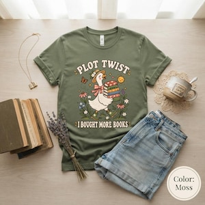 Könnte beinhalten: Ein olivgrünes T-Shirt mit einer Grafik einer weißen Gans, die einen Strohhut und grüne Stiefel trägt und einen Stapel bunter Bücher hält. Der Text lautet PLOT TWIST I BOUGHT MORE BOOKS, umgeben von Blumen, einem Schmetterling und einer Sonne.