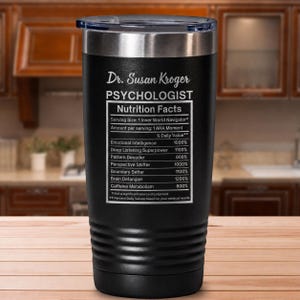 May include: A black stainless steel tumbler with a clear lid, featuring white text that reads Dr. Susan Kroger PSYCHOLOGIST Nutrition Facts, listing various attributes like Emotional Intelligence 1000% and Deep Listening Superpower 1100%.