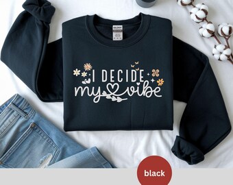 May include: Black crewneck sweatshirt with the text "I DECIDE my vibe" in white script, decorated with floral and butterfly graphics. The sweatshirt is displayed on a white surface with a pair of blue jeans.