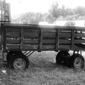 May include: A black and white image of an antique wooden wagon with four wheels. The wagon bed is constructed of horizontal wooden planks. The wheels are black with white rims. The wagon is parked on grass.