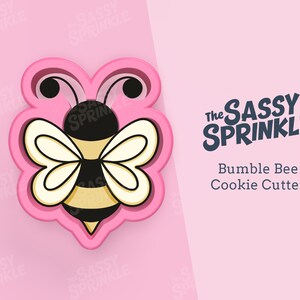 May include: Pink plastic cookie cutter shaped like a cartoon bumblebee with black eyes, yellow and black stripes, and white wings. The text "The SASSY SPRINKLE" and "Bumble Bee Cookie Cutter" is in the background.