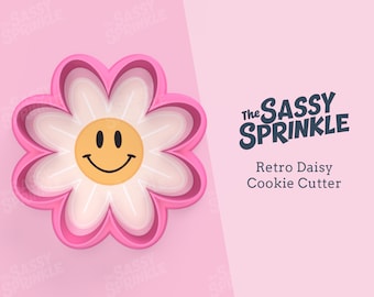 May include: Pink plastic cookie cutter shaped like a daisy with a yellow smiley face in the center. The text "The Sassy Sprinkle" and "Retro Daisy Cookie Cutter" is in the background.
