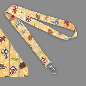 May include: A yellow lanyard with a repeating pattern of colorful snakes. The lanyard has a silver clip and is approximately 1.5 inches wide.