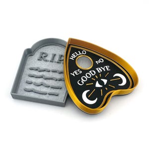 May include: A black and gold plastic cookie cutter shaped like a Ouija board with the words "Hello", "Yes", "No", "Goodbye" and a crescent moon and eye design. The cookie cutter is open and next to a gray plastic cookie cutter shaped like a tombstone with the word "RIP" on it.