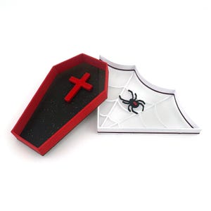 May include: A red coffin-shaped box with a black interior and a red cross on the lid. The box is open and the lid is resting on a white spiderweb with a black spider.
