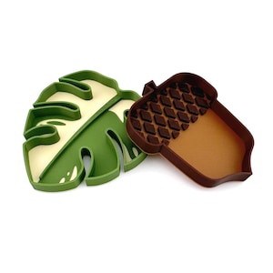 May include: Two green and brown silicone cookie cutters, one shaped like a monstera leaf and the other shaped like an acorn.