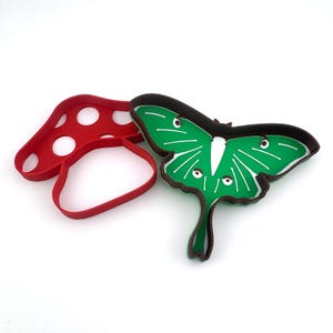 May include: Two cookie cutters, one shaped like a red mushroom and the other shaped like a green luna moth.