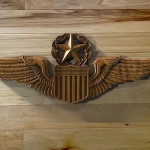 May include: A wooden carved pilot wings badge with a shield and a star in a wreath. The wings are spread out and the shield is in the center. The badge is on a wood background.