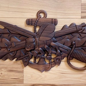 May include: A wooden carving of a Navy SEAL insignia. The insignia features an anchor, a rifle, a pistol, a knife, a sword, and oak leaves. The carving is dark brown and has a detailed design.
