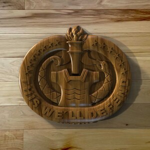 May include: A wooden carving of the United States Army's crest. The crest features a torch, a shield, and two eagles. The text "This We'll Defend" is inscribed around the crest.