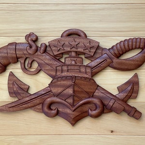 May include: A carved wooden emblem featuring a ship, anchor, sword, and pistol, with three stars on a banner. The dark brown wood carving is mounted on a light wood background.