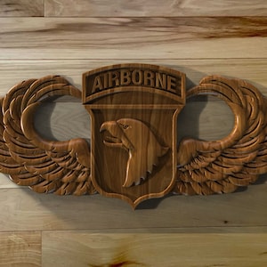 May include: A wooden Airborne badge with a detailed eagle emblem and the word "AIRBORNE" carved above. The badge features stylized wings on either side and is set against a wooden background.