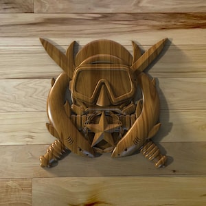 May include: A wooden carving of a scuba diver's mask with crossed knives and two sharks on either side. The carving is on a wooden background.