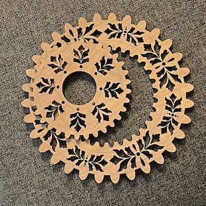 May include: Two wooden gear-shaped decorative pieces with intricate leaf and floral cutouts. The gears are light brown with dark brown details, creating a contrast. The design is symmetrical and detailed, suitable for home decor or craft projects.