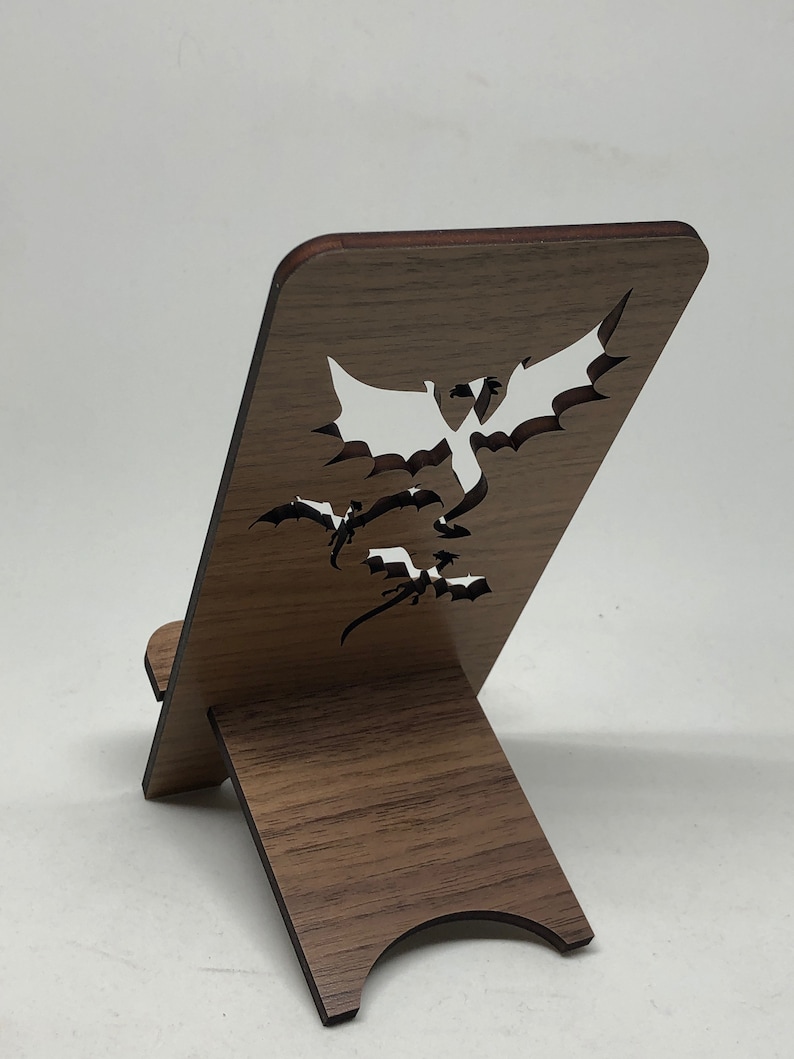 May include: A wooden phone stand with a dragon design cut out. The stand is brown with a dark brown trim. The stand is folded up and ready to use.