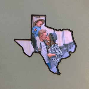 May include: A family portrait in a Texas-shaped frame. A couple is kissing, and a child is sitting on the man's shoulders. The photo is in color and has a vintage feel.