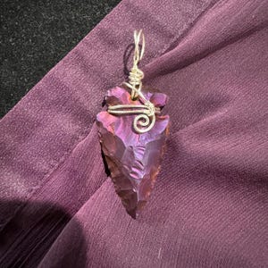May include: A purple arrowhead pendant wrapped in silver-colored wire. The stone has a translucent, glossy finish with visible flaked edges. The wire forms a decorative spiral detail at the center and a loop at the top for hanging the pendant.