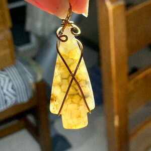 May include: A gold wire wrapped pendant featuring a light brown agate stone with a unique pattern. The stone is shaped like a teardrop and has a wire loop at the top for hanging.