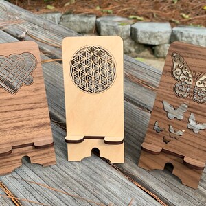 May include: Three wooden phone stands with different laser cut designs. The first stand has a Celtic knot heart design, the second has a flower of life design, and the third has a butterfly design.