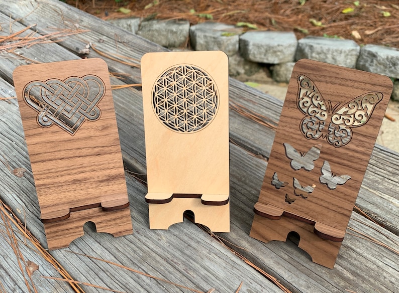 May include: Three wooden phone stands with different laser cut designs. The first stand has a Celtic knot heart design, the second has a flower of life design, and the third has a butterfly design.