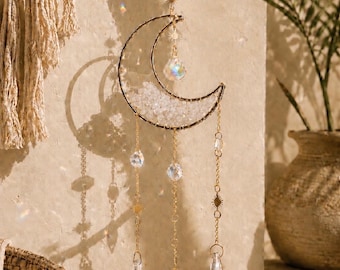 May include: A gold-toned crescent moon suncatcher featuring a wire-wrapped frame filled with clear, textured glass fragments. Three gold chains hang from the moon, each adorned with clear, faceted crystal beads and pointed crystal pendants.