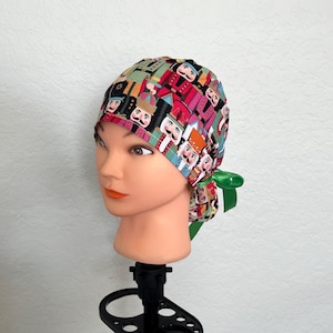 May include: A colourful scrub cap featuring a repeating pattern of nutcracker figures in various colours. The cap is tied in the back with a green ribbon. The cap is displayed on a mannequin head.