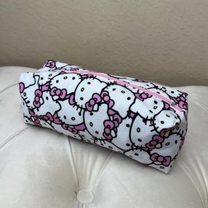 May include: A rectangular pencil case featuring a repeating pattern of white Hello Kitty faces with pink bows on a white background. The case has a pink zipper closure.