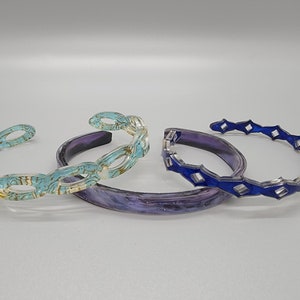 Puede incluir: Tres pulseras de resina. Una es azul con un patrón de diamante, otra es turquesa con un patrón dorado y la última es morada con un patrón de galaxia.